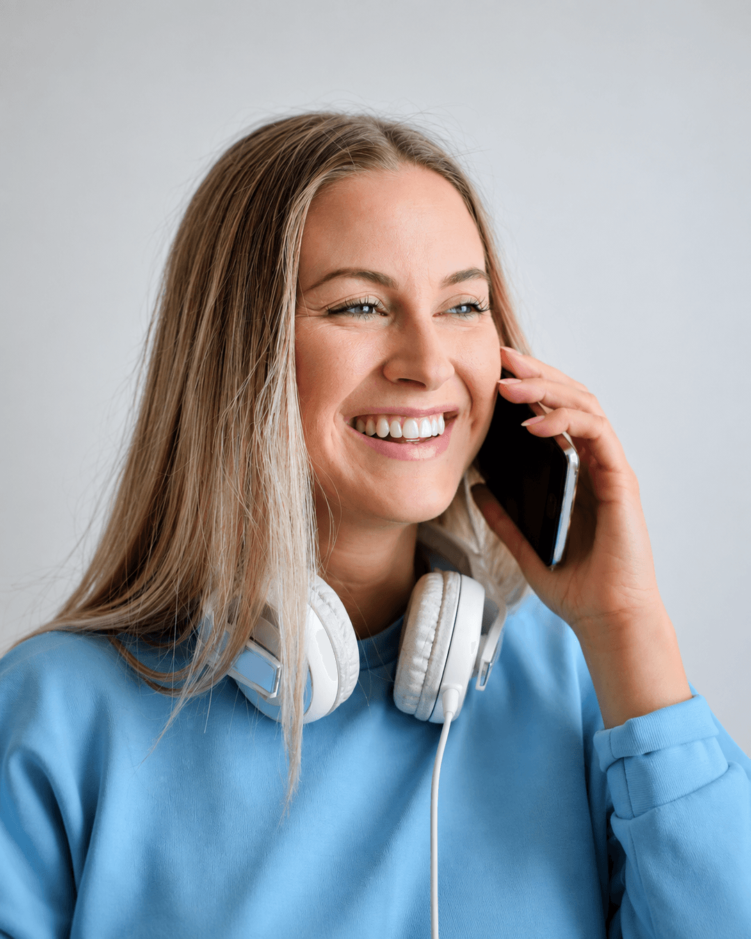 A smiling woman in a blue top talking on a smartphone, with headphones around her neck.